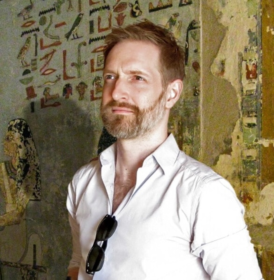A person standing in front of an ancient Egyptian wall with hieroglyphics and artwork. The person's face is blurred, and they are wearing a light-coloured shirt with sunglasses hanging from the collar.